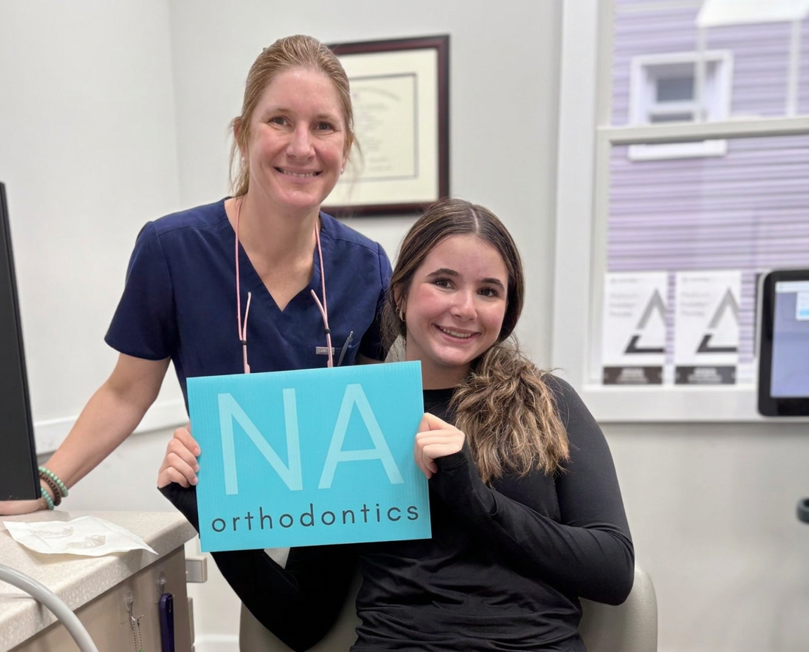 Patient and doctor smiling with the NA Orthodontics sign inside the office